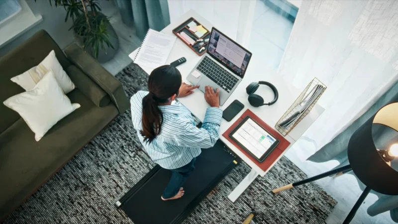 Frau geht auf einem Walking Pad unter dem Schreibtisch und arbeitet dabei am Laptop
